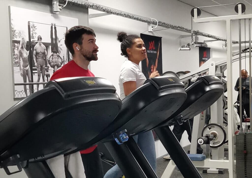Twee sporters – een man en een vrouw – lopen naast elkaar op loopbanden. De drie loopbanden staan in een rij, met een energieke oldschool sfeer op de achtergrond."